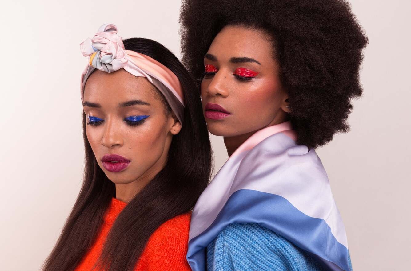 Makeup by Lucia Pilin on two melanated models. They are both wearing graphic eyeliner one in red and the other in electriv blue, glossed up.